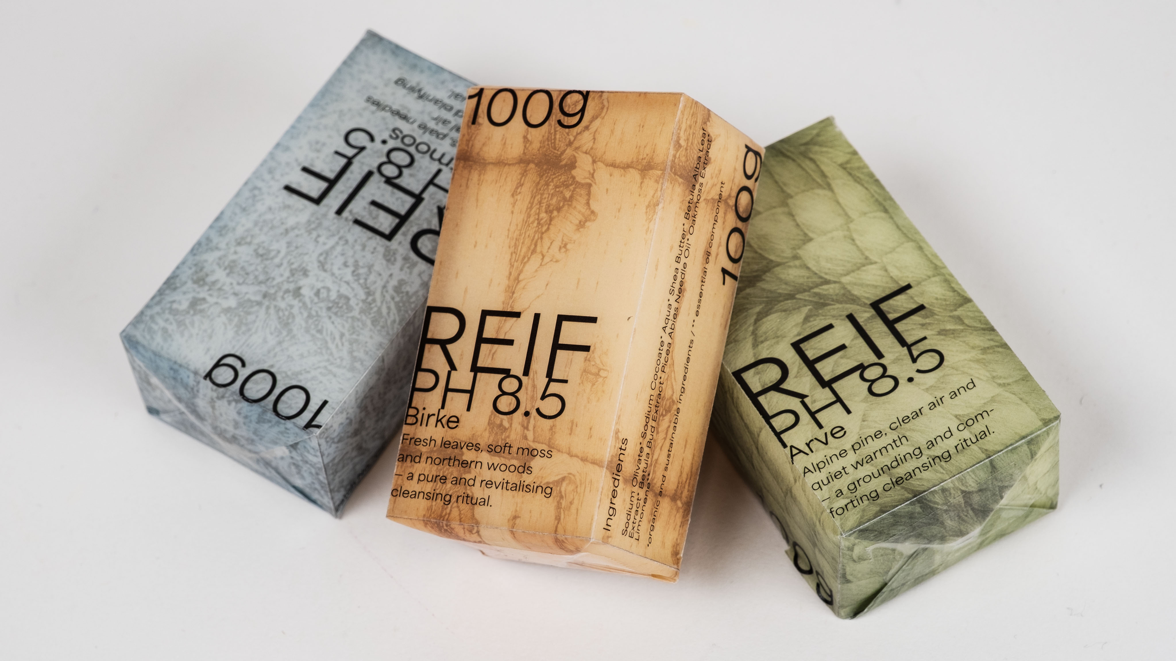 Three rectangular bars of REIF PH8.5 soap in textured packaging featuring natural themes of blue moss, beige birch wood, and green pine.