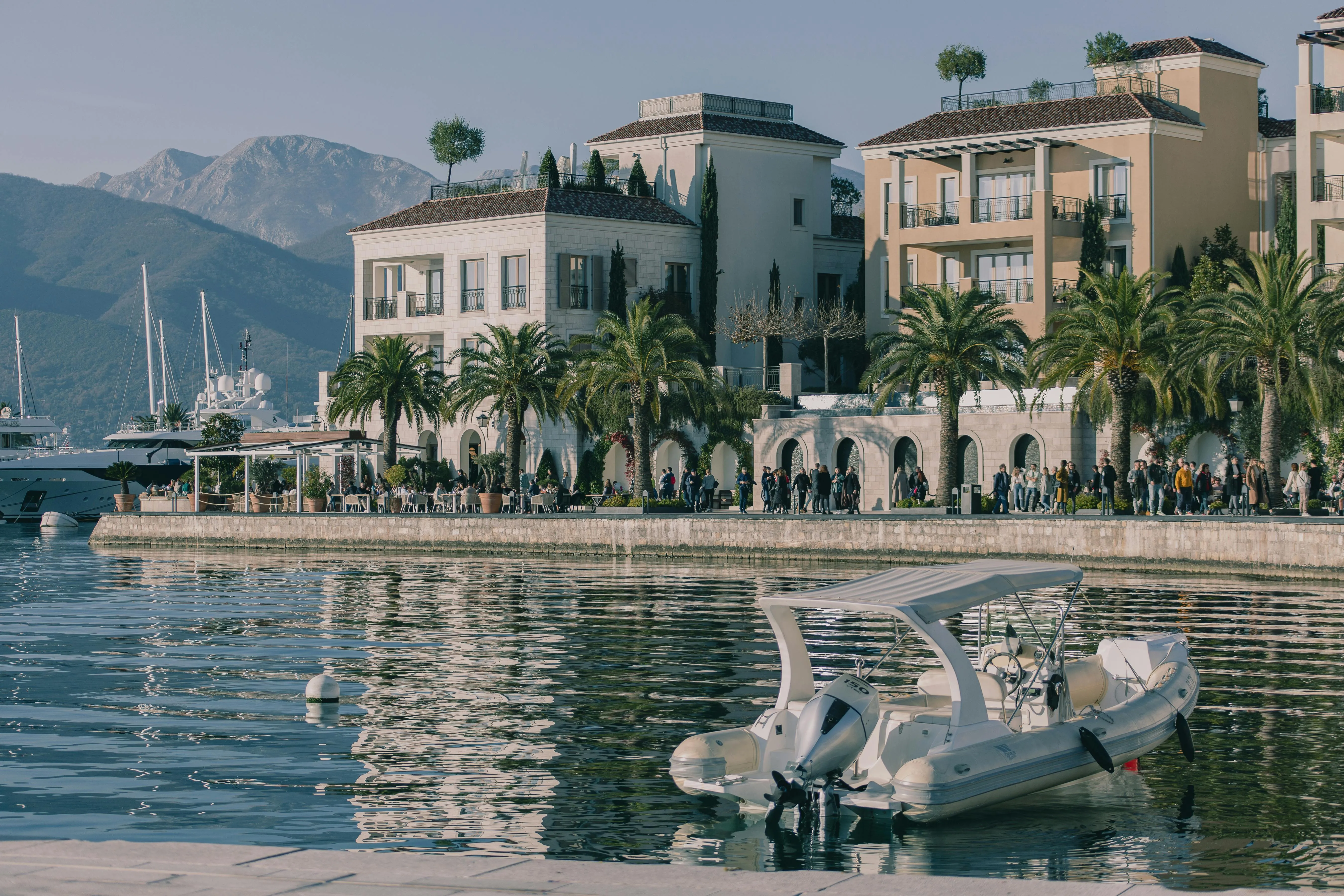 Small white motorboat moored in calm waterfront with palm trees, people, and buildings against mountainous background.