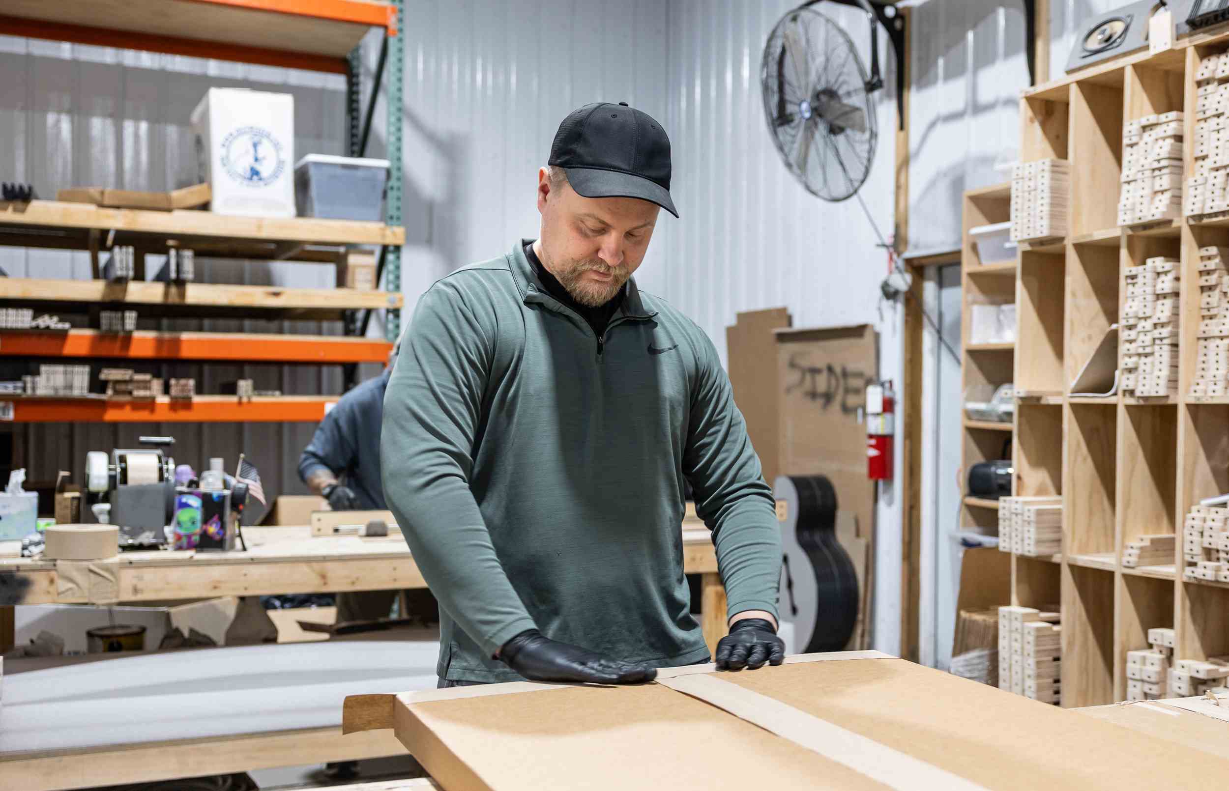 Man wearing black gloves and cap packaging a large cardboard box in a workshop with wooden shelves and tools.