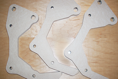 Three identical beige engine mounting brackets with multiple holes placed overlapping on a wooden surface.