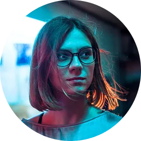 Portrait of a woman with short hair and glasses illuminated by blue and orange lighting.