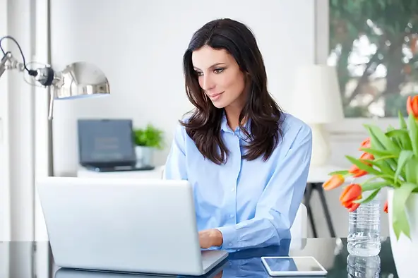 Dedicated virtual assistant USA focused on laptop tasks in a bright home office with flowers and a tablet.