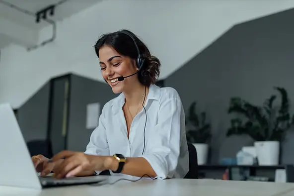 Business virtual assistant services professional smiling while using a headset and typing on a laptop.