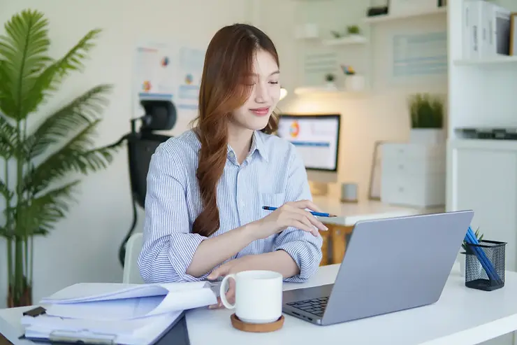 Customer service virtual assistant working remotely with documents, coffee, and a laptop in a bright office space.