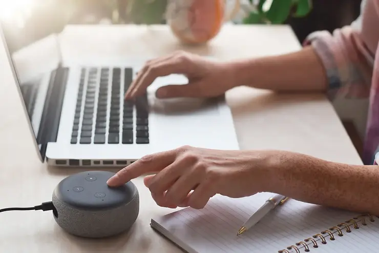 Virtual assistant setup with a person tapping a smart speaker while typing on a laptop at a home office desk.