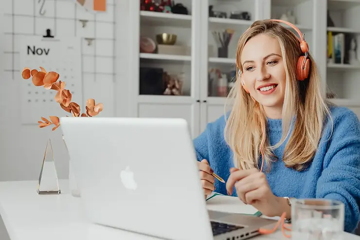 Remote virtual assistant smiling and taking notes on a MacBook while wearing orange headphones in a bright home office.