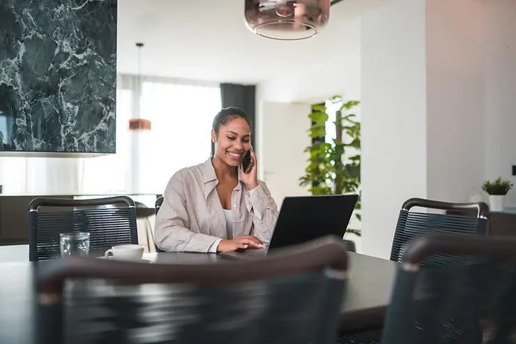 Remote virtual assistant smiling while talking on the phone and typing on a laptop at a modern home dining table.