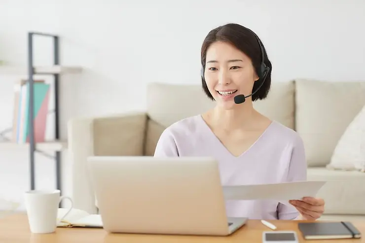 Remote virtual assistant smiling in a headset while reviewing a document beside a laptop in a cozy home office.