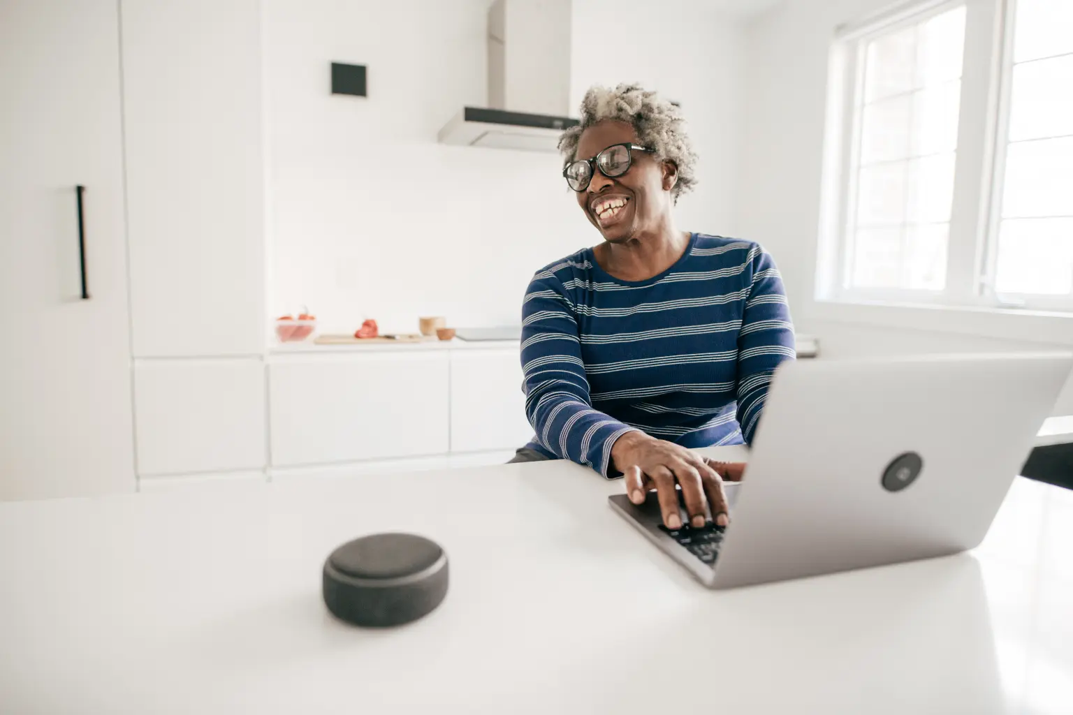 Virtual assistant services made easy as a woman laughs while typing on a laptop beside a smart speaker in a minimal home kitchen.