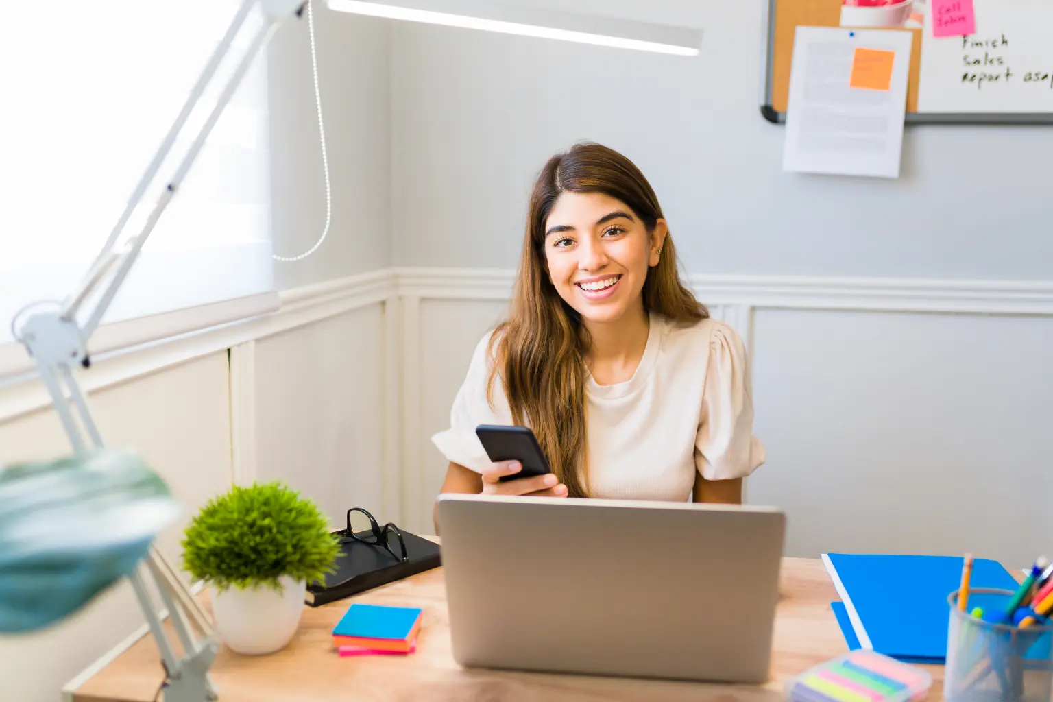 Virtual assistant smiling at her desk while holding a smartphone beside a laptop in a bright, organized home office.
