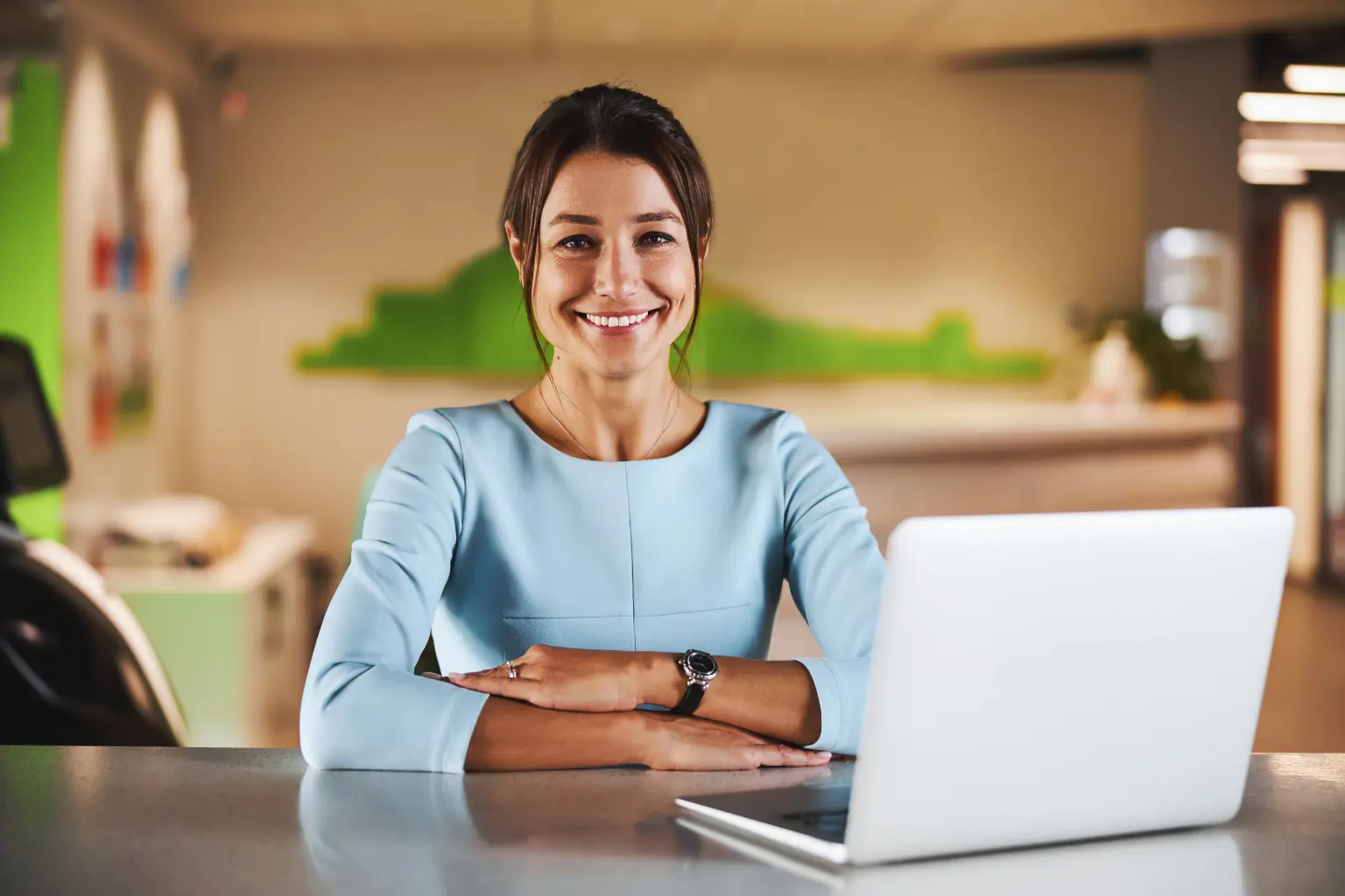 Virtual receptionist smiling confidently at her desk with a laptop open in a modern, bright office environment.