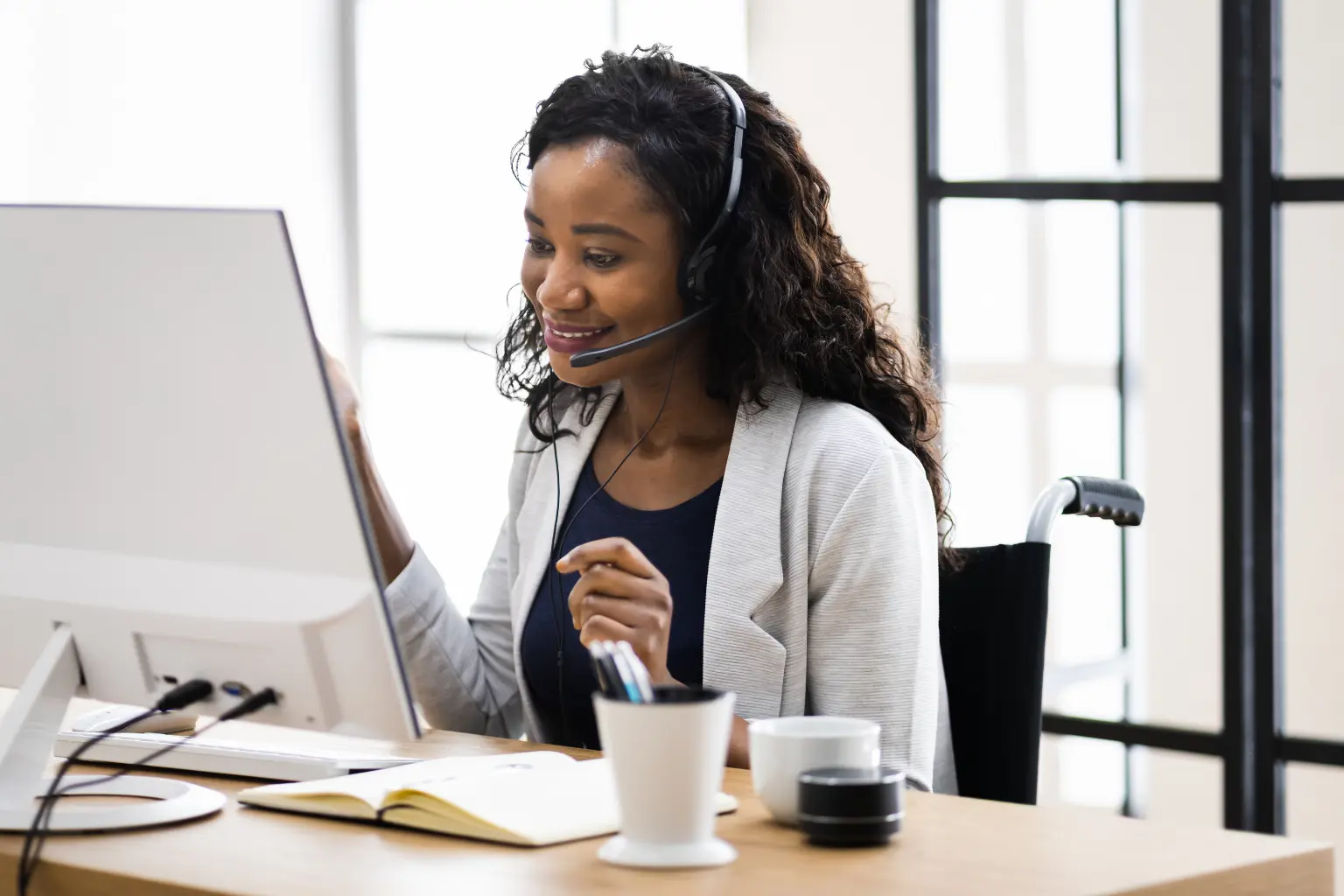 Remote VA in a wheelchair wearing a headset and adjusting a monitor while taking notes at a well-lit office desk.