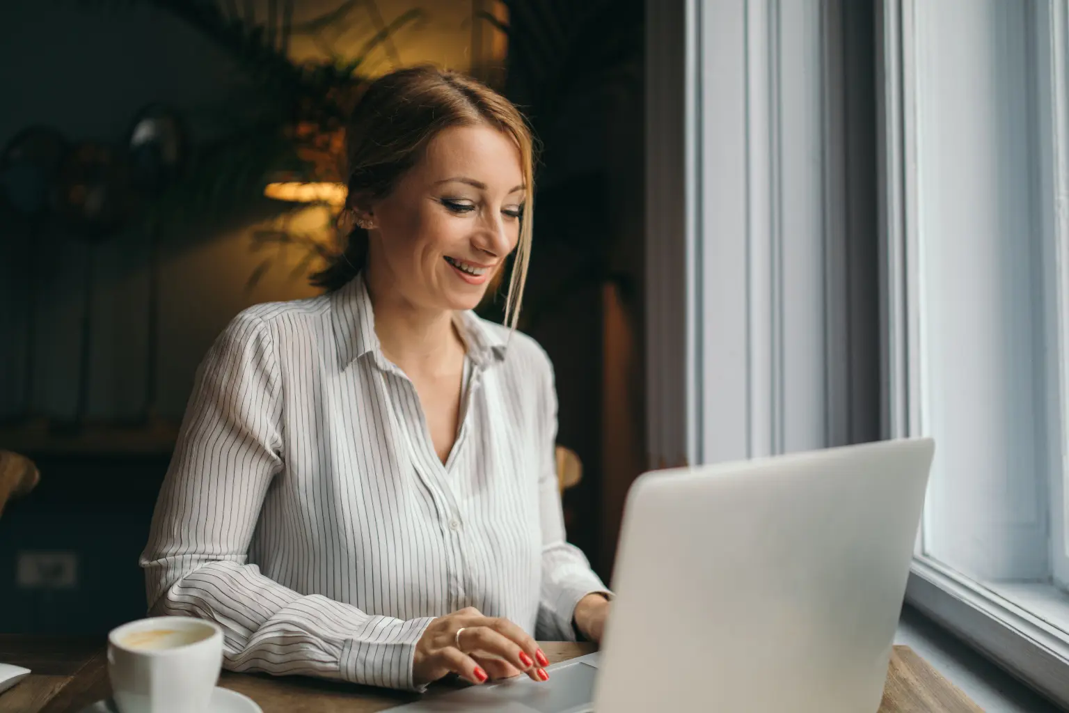 Virtual assistant smiling while typing on a laptop beside a coffee cup at a window-side café table.