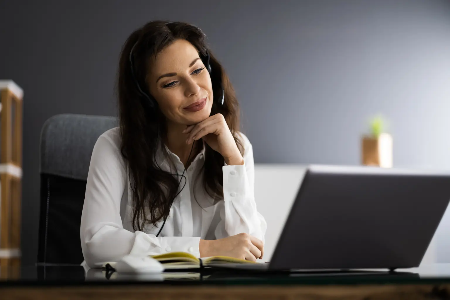 Remote receptionist wearing a headset and resting her chin thoughtfully while reviewing a laptop screen and open notebook.