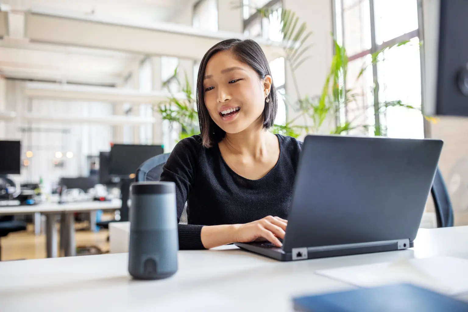 Virtual assistant smiling and typing on a laptop beside a smart speaker in a sunlit open-plan office.