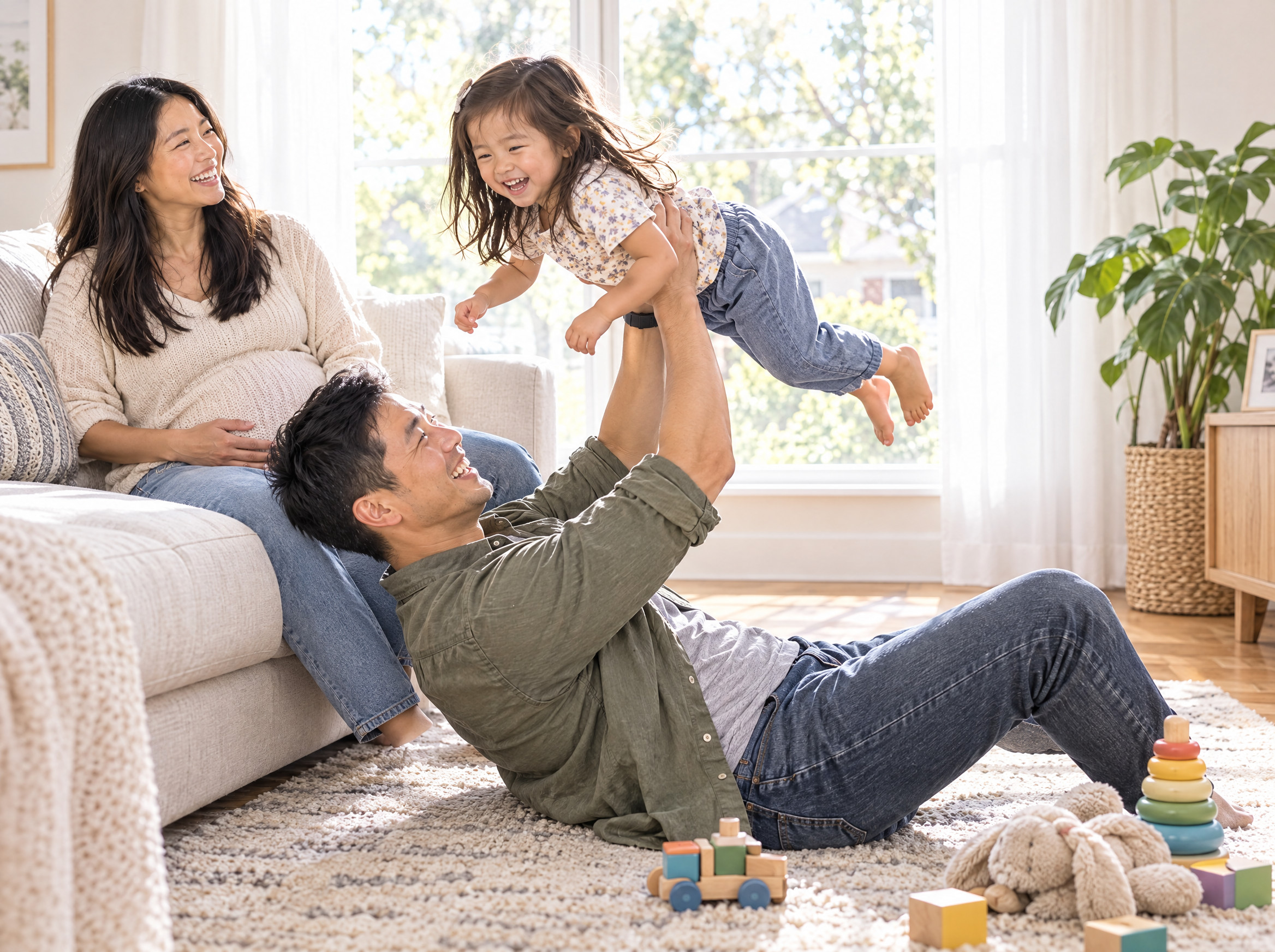 A young family playing in a bright living room, dad lifting their toddler overhead while mom, pregnant, laughs from the sofa
