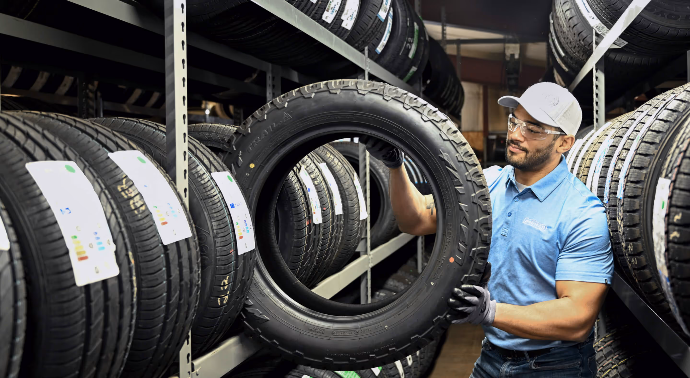 Man wearing safety glasses, gloves, and a cap holding a large tire in a warehouse filled with rows of tires.