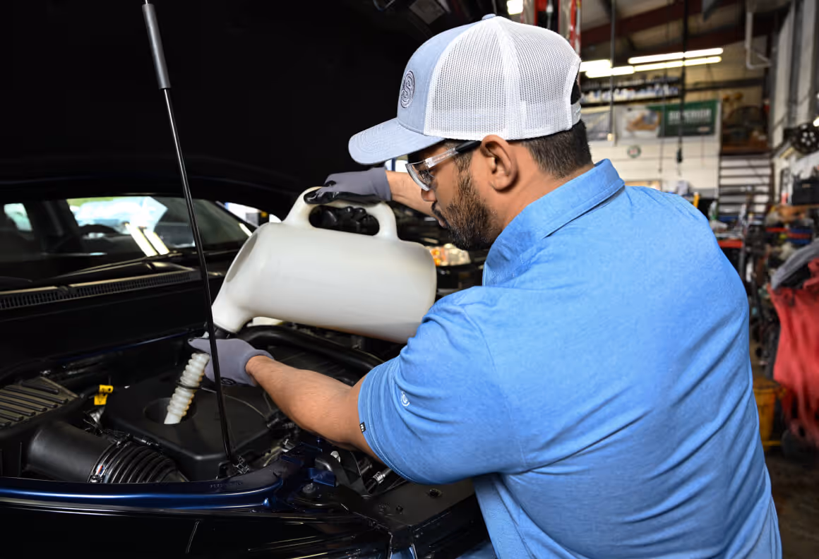 Man wearing a blue shirt and white cap pouring fluid into a car engine in a garage.