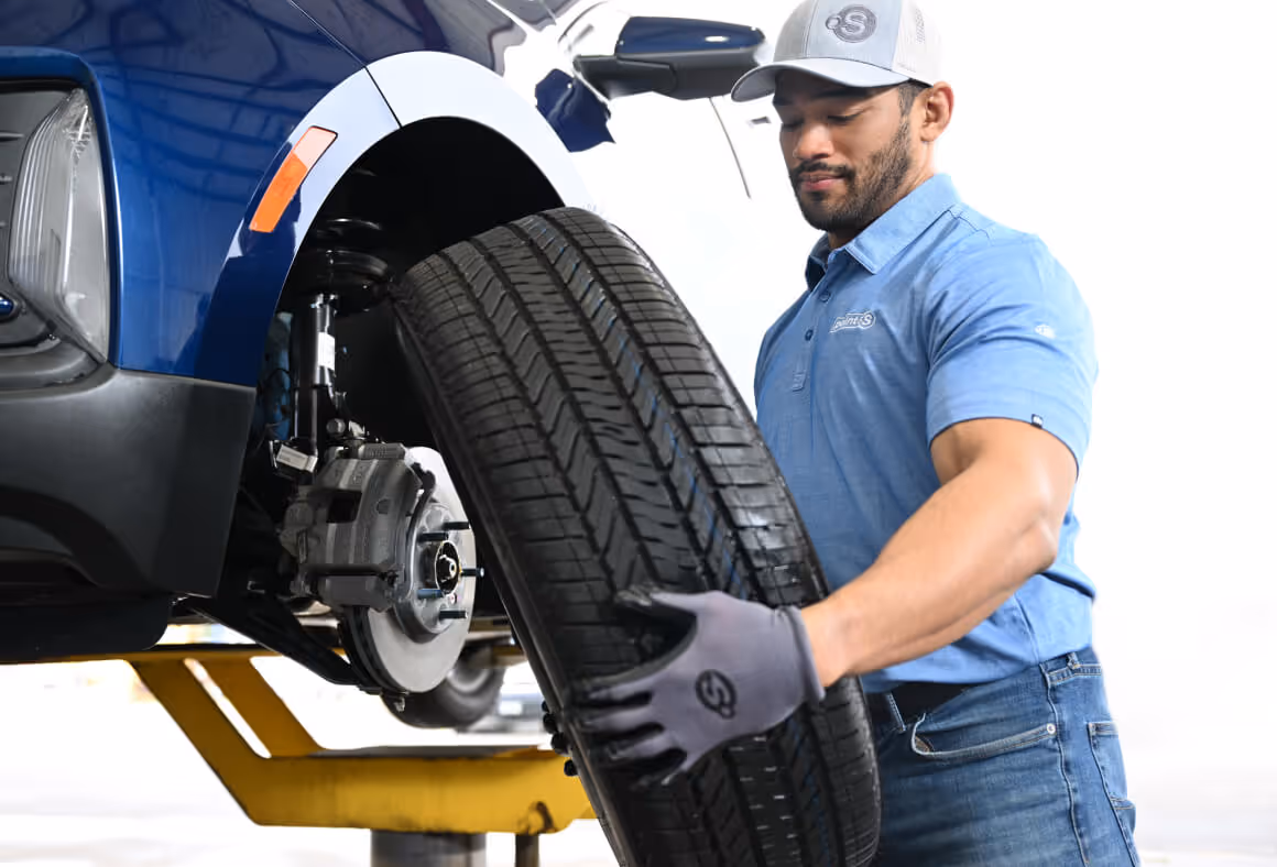 Mechanic in blue shirt and gloves holding a tire next to a blue vehicle lifted on a hydraulic lift.