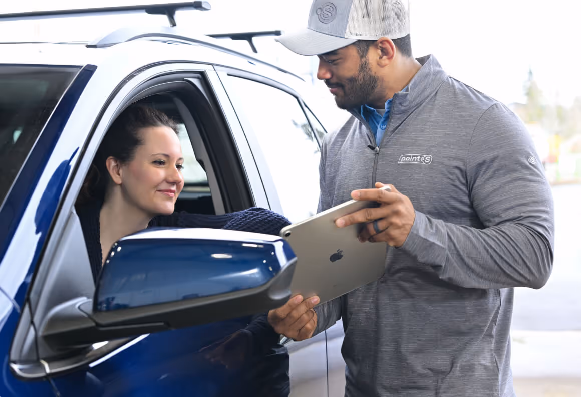 Man in a gray jacket and cap holding a tablet, assisting a woman seated in a blue car.