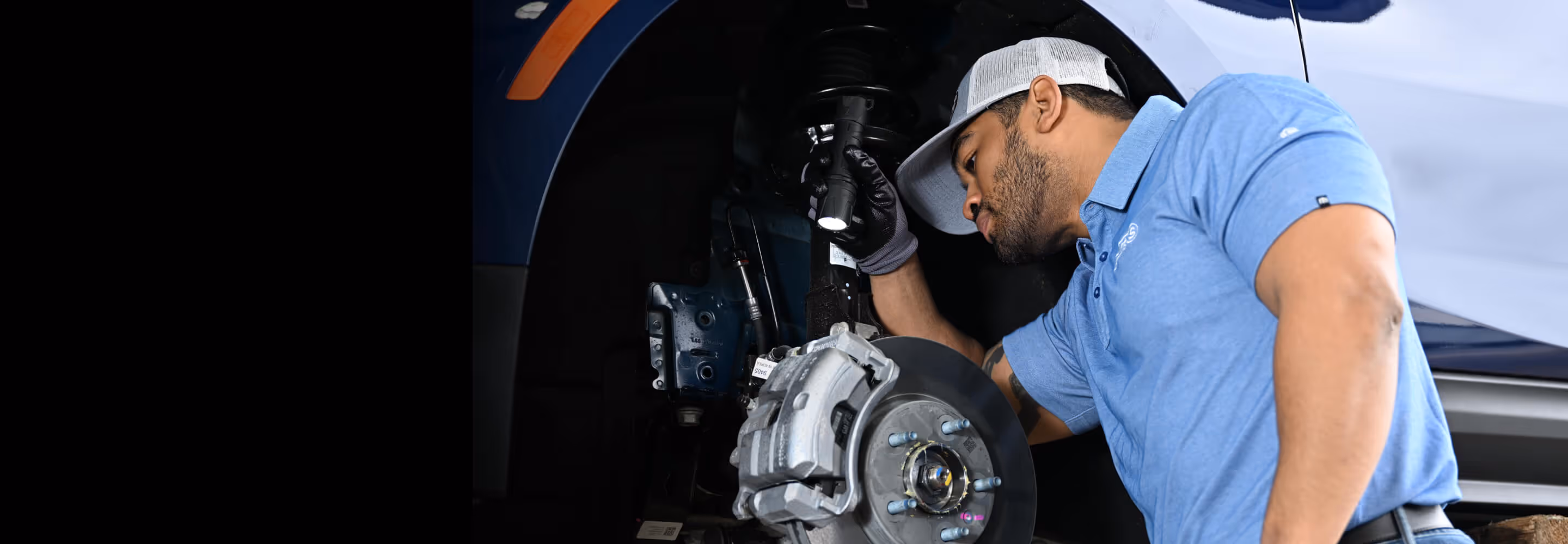 Mechanic in blue shirt and cap inspecting a car brake system with a flashlight.