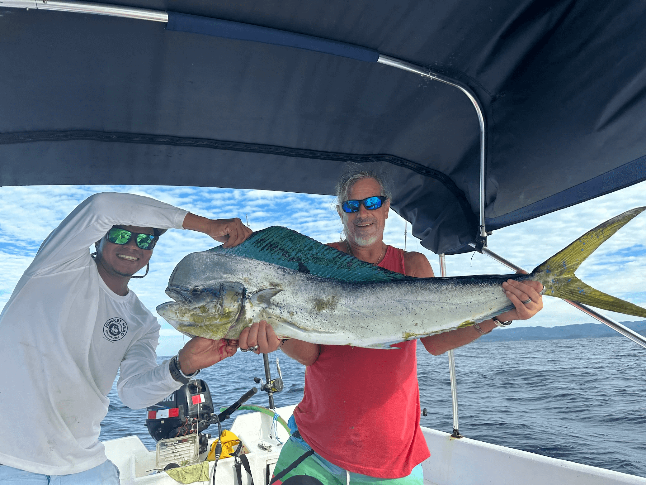Two men on a boat holding a large mahi-mahi fish with ocean and partly cloudy sky in the background.