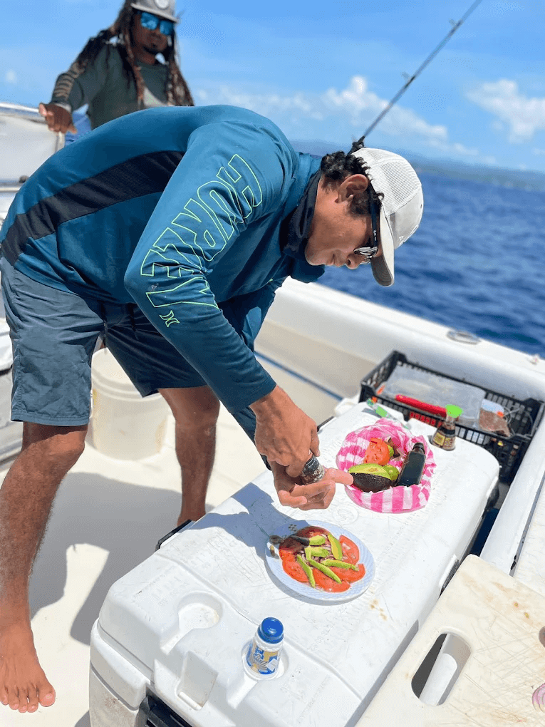 Man in a white cap and blue long-sleeve shirt preparing food with avocado and tomato on a boat with ocean in the background.