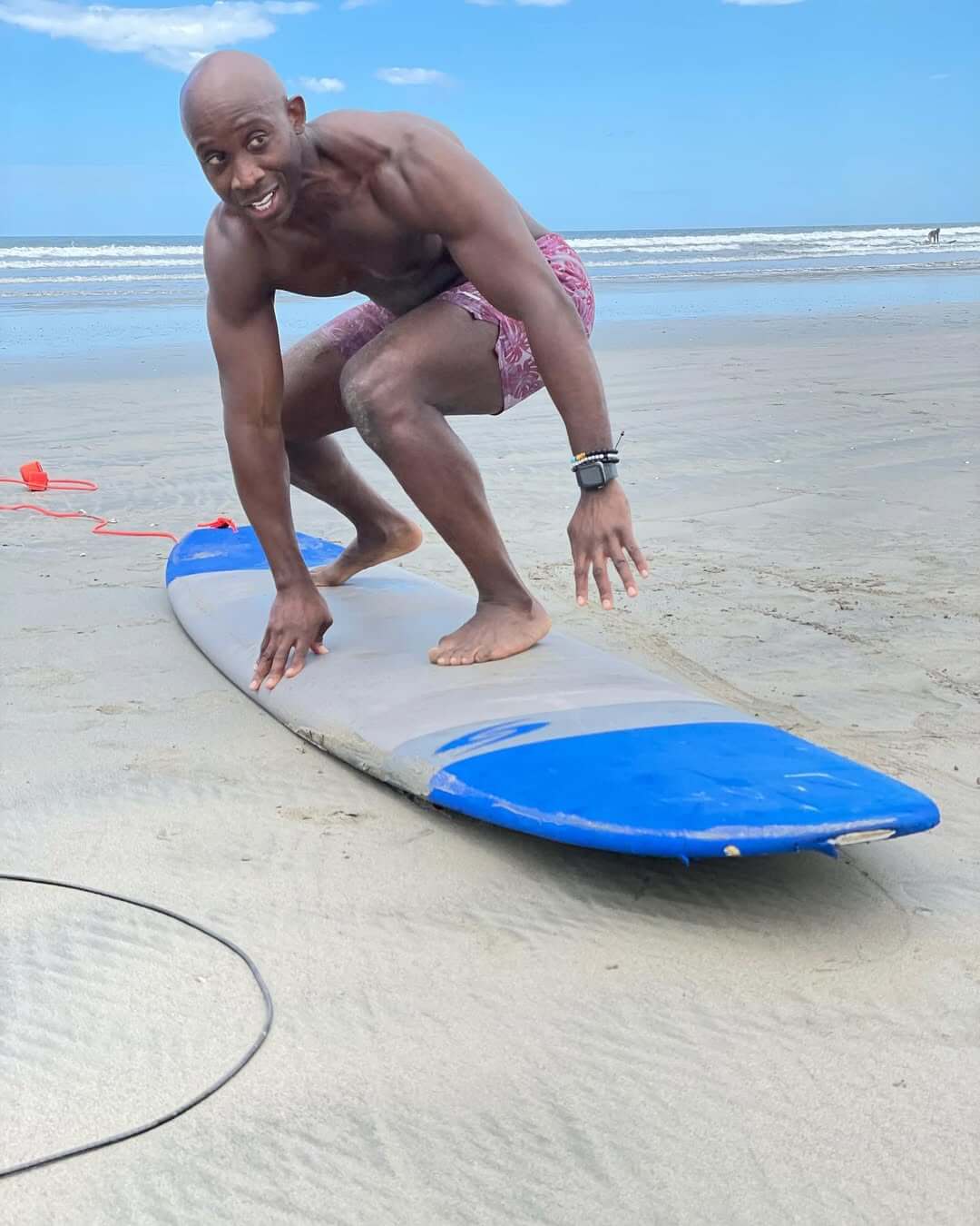 Man in pink shorts crouching on a surfboard on the sandy beach near the ocean.