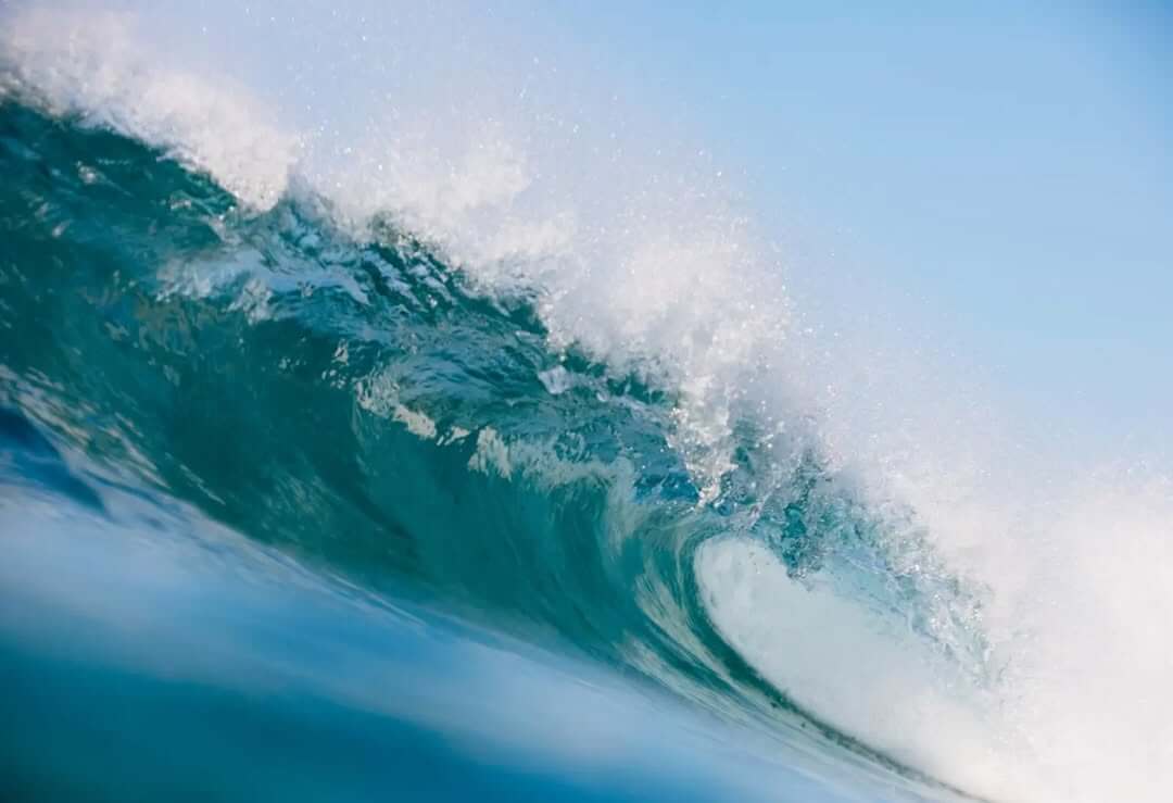 Close-up of a breaking ocean wave with clear blue water and white foam under a bright sky.