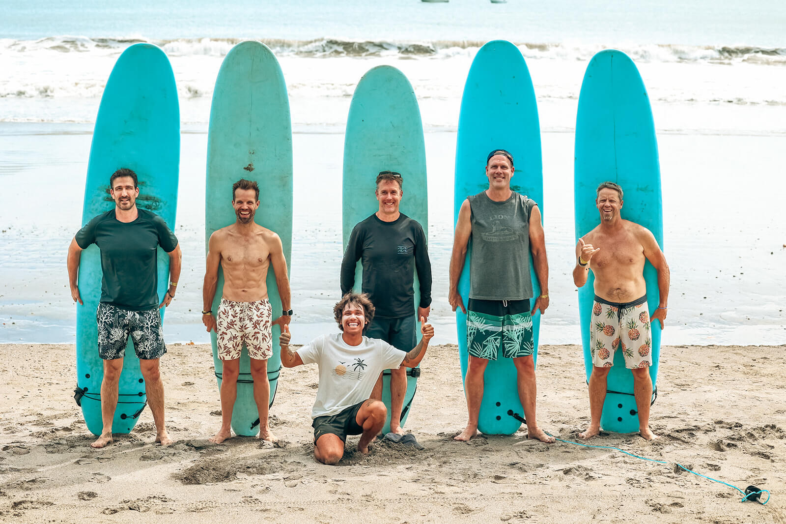 Five men standing on a sandy beach holding blue surfboards upright behind them, with one man kneeling in front showing thumbs-up gestures, ocean waves in the background.