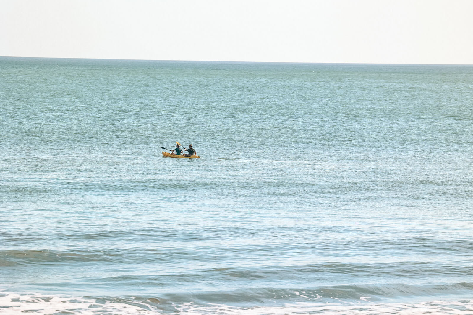 Two people kayaking together on calm ocean water under a bright sky.