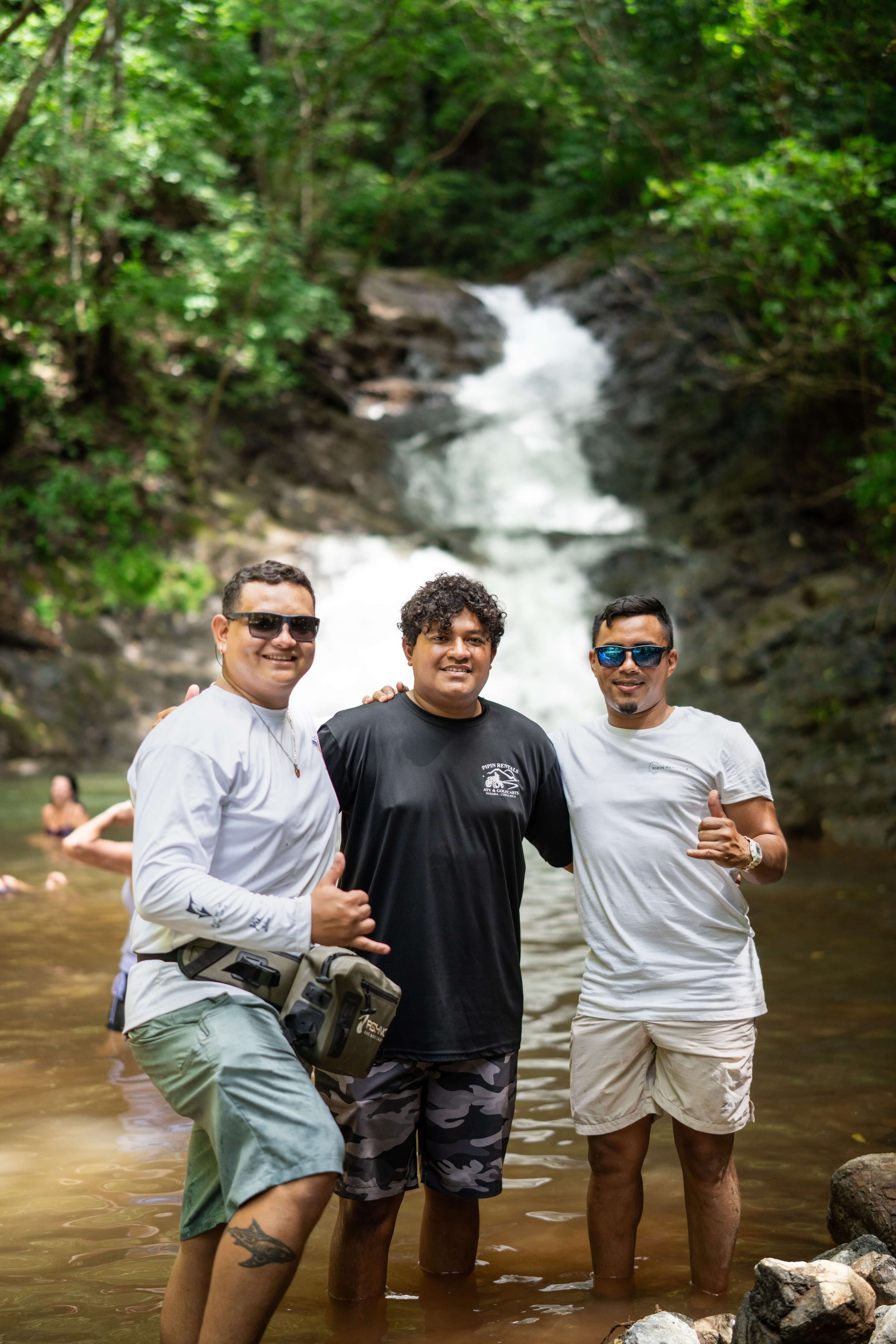 Three men standing in shallow water with a waterfall and green forest in the background, smiling and making shaka hand signs.