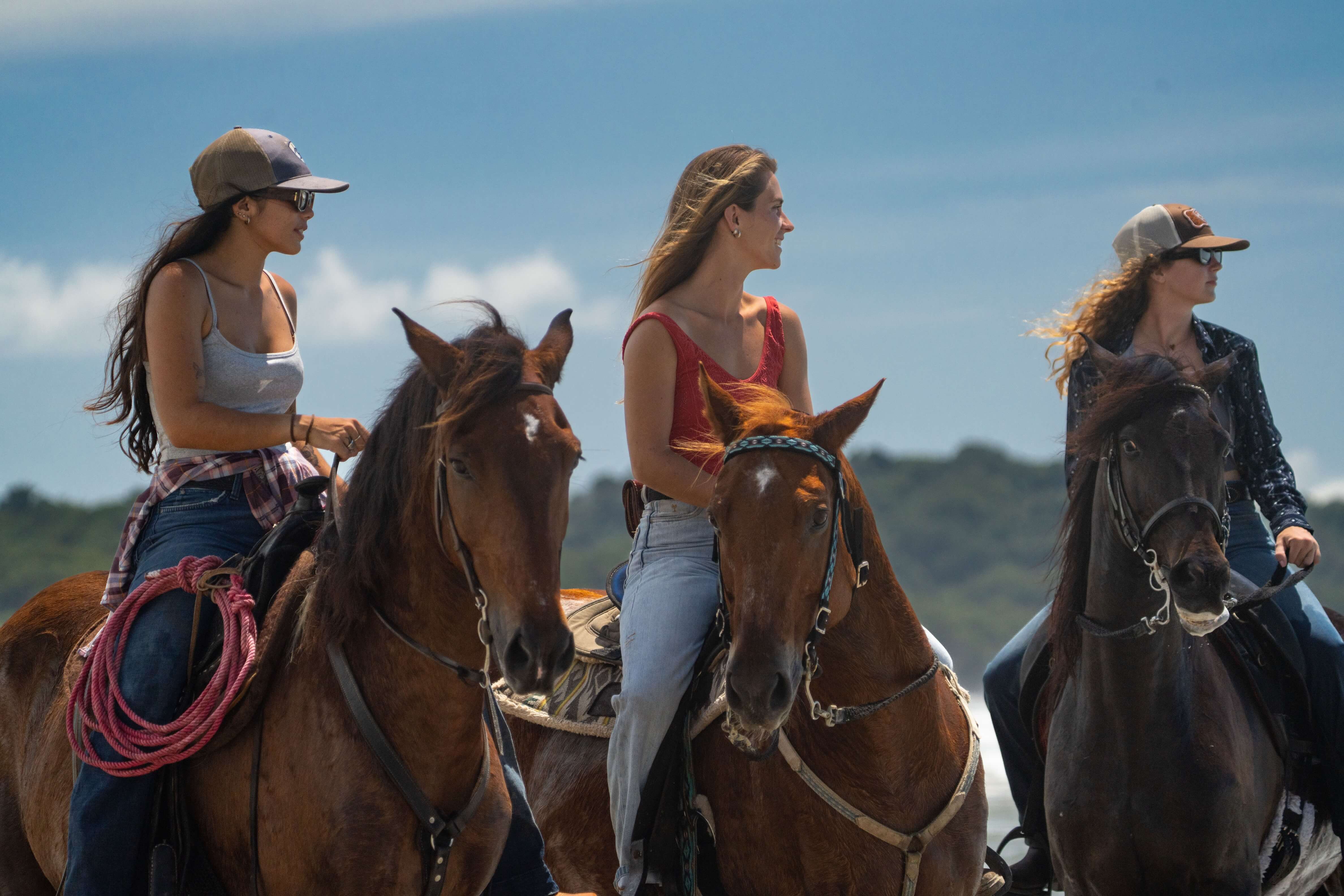 Three women riding horses outdoors on a sunny day with a hill in the background.