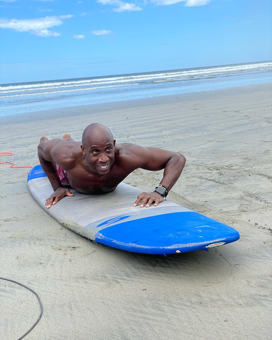 Man lying on a surfboard on the sandy beach practicing paddling with ocean waves in the background.