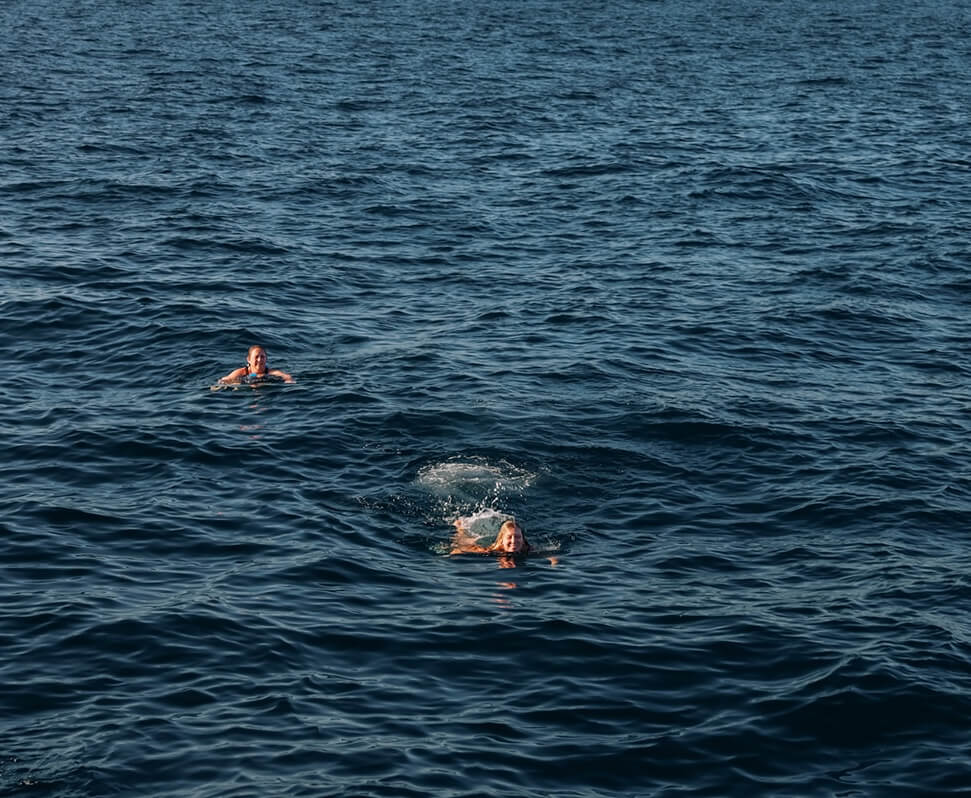 Aerial view of a rocky islet with a small sandy beach surrounded by turquoise ocean waves and a green, forested coastline in the background.