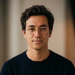 Portrait of a young man with short dark hair, wearing round glasses and a black shirt, looking at the camera with a neutral expression.