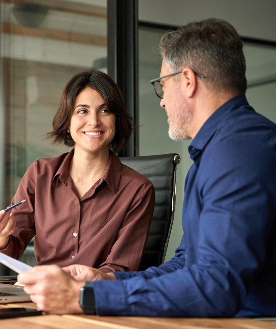 A smiling woman in a brown shirt holds a pen and talks with a man in a blue shirt during a meeting in an office.