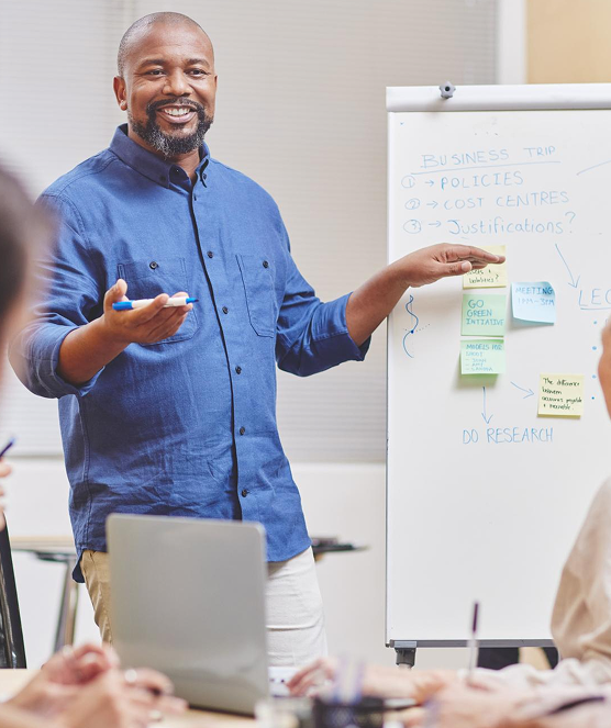 Man in a blue shirt giving a presentation and pointing to notes on a whiteboard during a meeting.