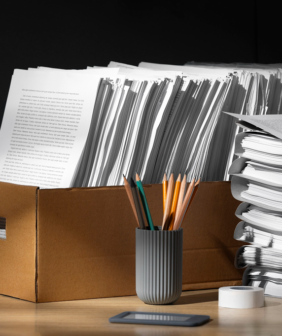 Stack of papers organized in a cardboard box and piles on a desk with pencils in a gray holder, a roll of tape, and a smartphone.