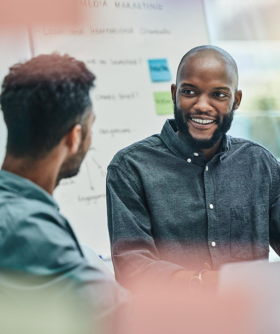 Two men having a discussion in an office with a whiteboard in the background containing notes about media marketing.