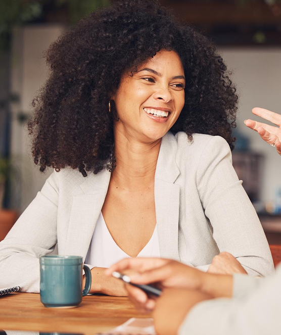 Smiling woman with curly hair wearing a light blazer, sitting at a table with a blue coffee mug and engaged in conversation.