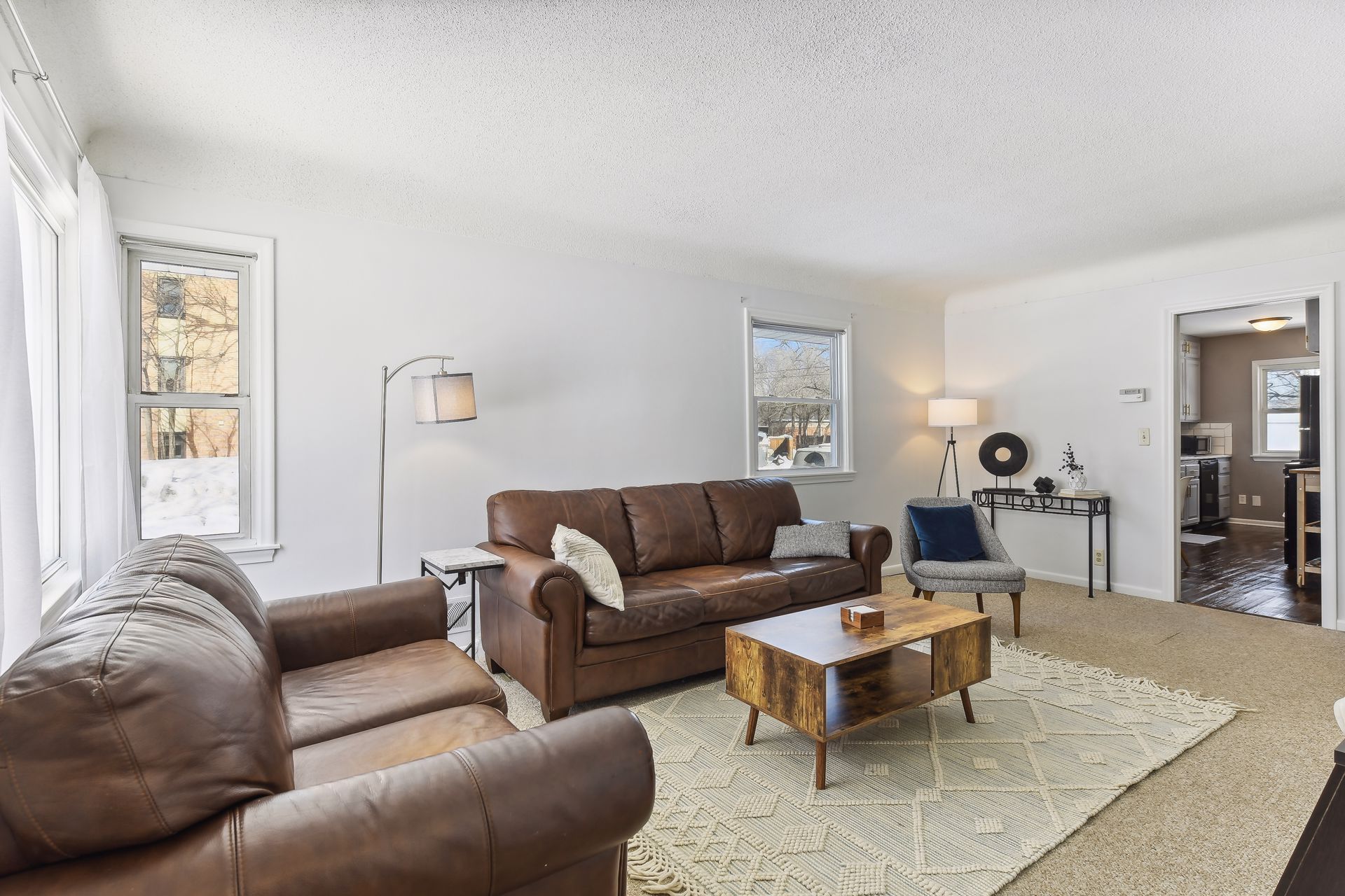 Living room with two brown leather sofas, a gray armchair, wooden coffee table on patterned rug, and floor lamp beside window.
