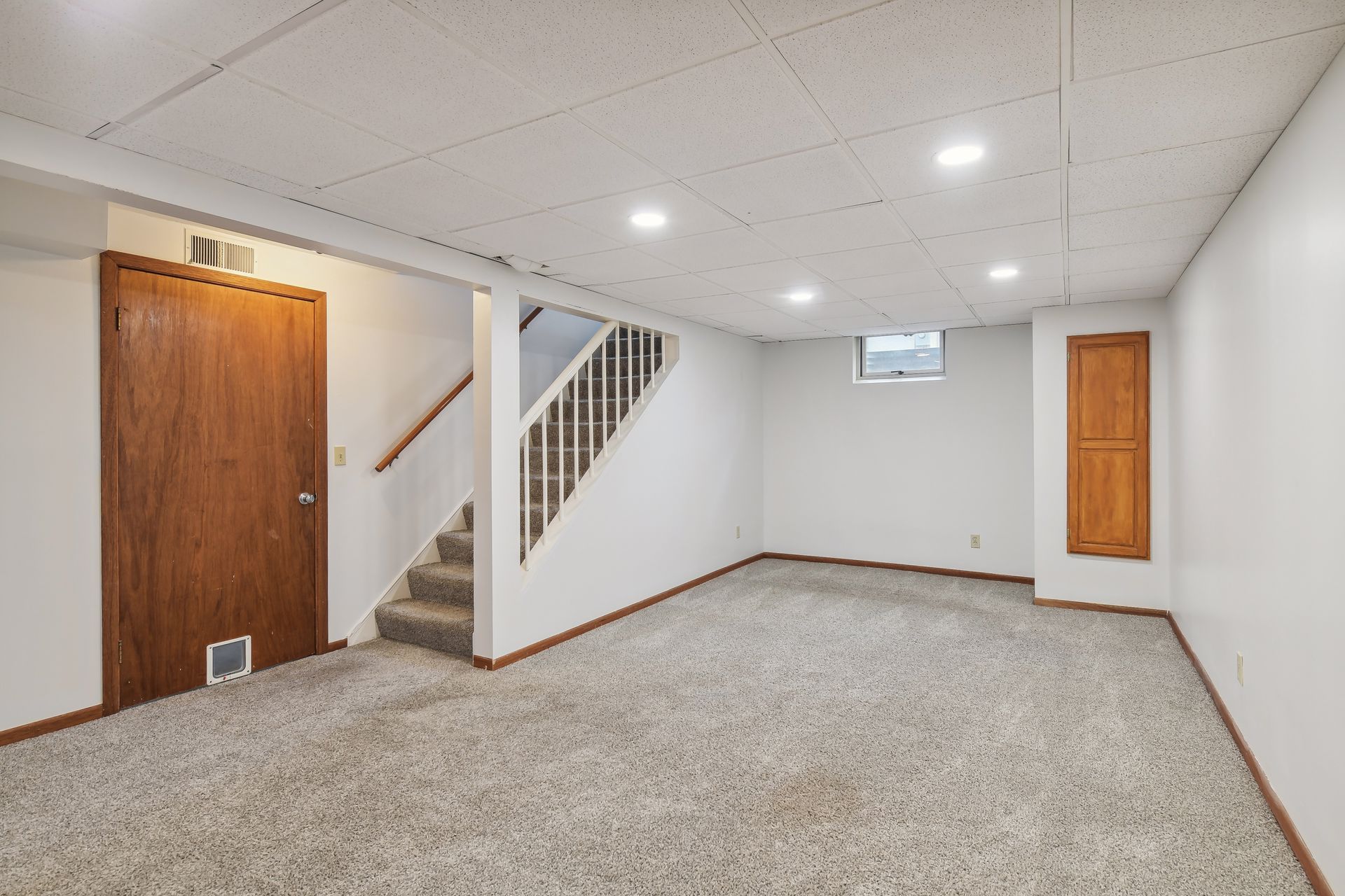 Empty basement room with beige carpet, white walls, wooden doors, stairs with railing, and a small window near the ceiling.