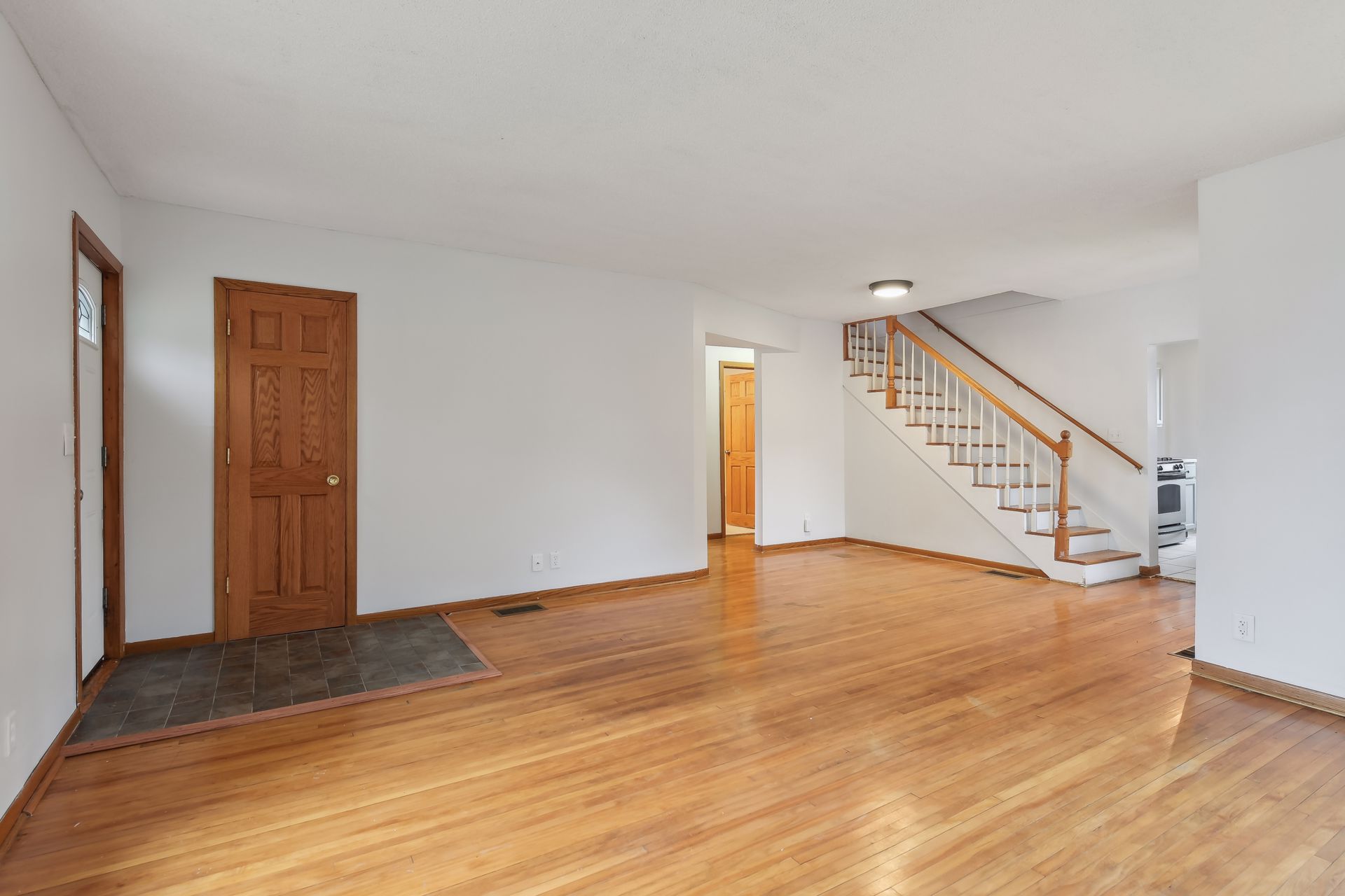 Empty living room with wooden floor, white walls, wooden staircase, and two wooden doors.