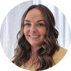 Smiling woman with long wavy brown hair wearing a yellow top in front of a white curtain background.