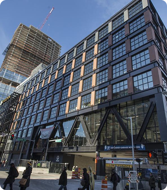Modern office building with large glass windows and an under-construction high-rise in the background on a city street with pedestrians.