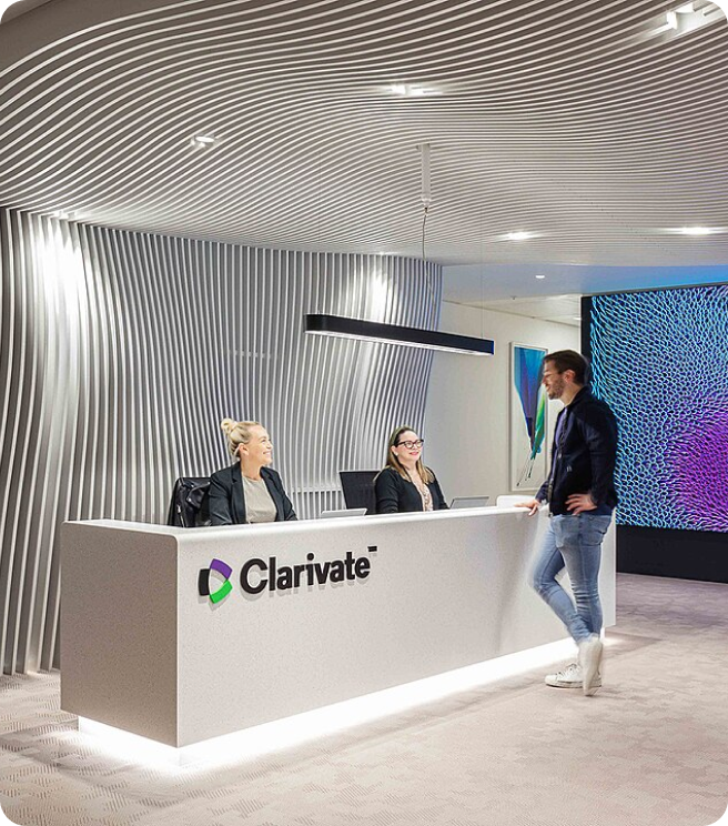 Modern Clarivate reception area with two seated women behind the desk and a man standing in front, featuring a curved white textured wall and colorful digital screen.