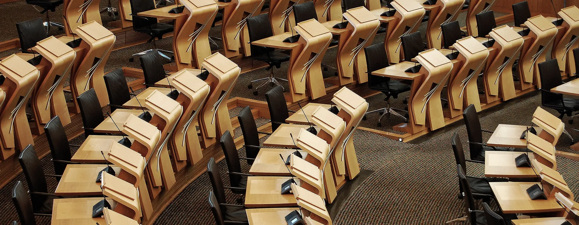 Rows of modern wooden desks with black chairs and microphones arranged in a semi-circular parliamentary chamber.