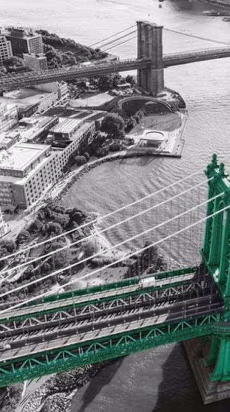 Aerial view of two riverside suspension bridges with the closest bridge highlighted in green against a black and white background.