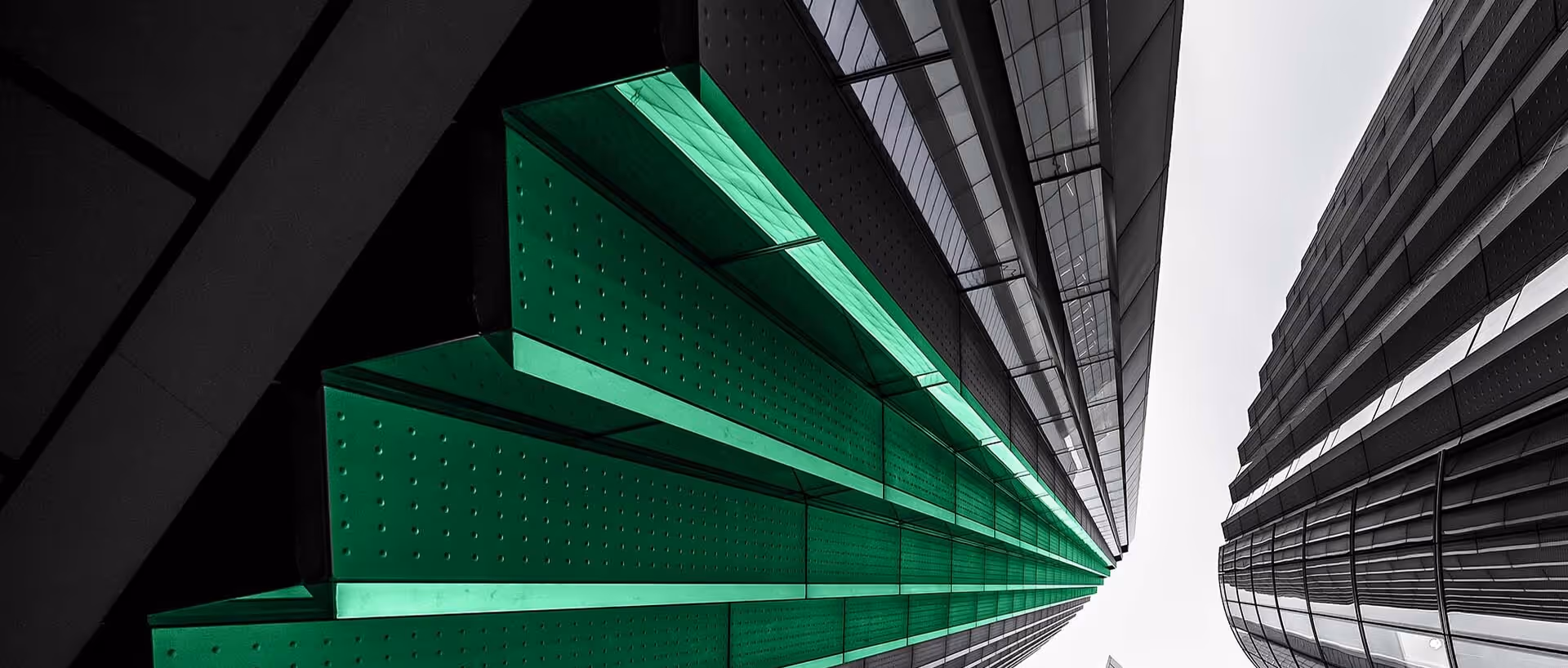 Upward view of tall modern buildings with green-lit panels on one side against an overcast sky.